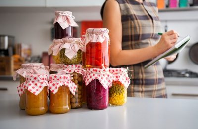 In the foreground, a stack of glass jars filled with home-made preserved vegetables. In the background, the profile of a woman listing the different ingredients before she stores them away for winter.