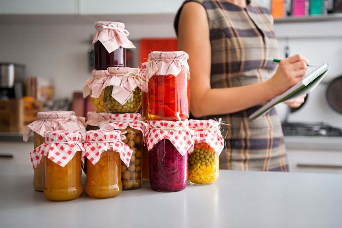 In the foreground, a stack of glass jars filled with home-made preserved vegetables. In the background, the profile of a woman listing the different ingredients before she stores them away for winter.